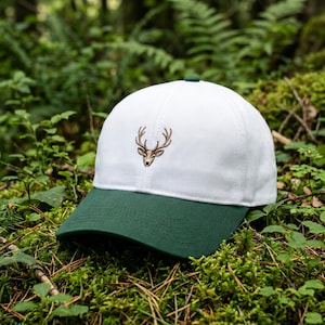 May include: A white baseball cap with a green peak and button. The cap has an embroidered brown deer head design. The hat is set against a backdrop of green foliage.