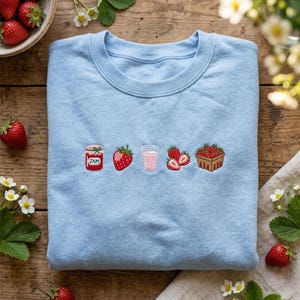 May include: Light blue sweatshirt featuring embroidered strawberry-themed designs. The design includes a jar of jam, a strawberry, a glass of pink drink, two strawberries, and a basket of strawberries. The sweatshirt is made of a soft material.
