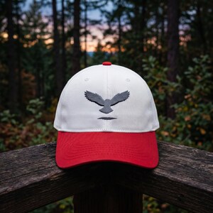 May include: A white and red baseball cap with an embroidered gray owl design. The cap has a red brim and a red button on top. The background is a blurred forest scene.