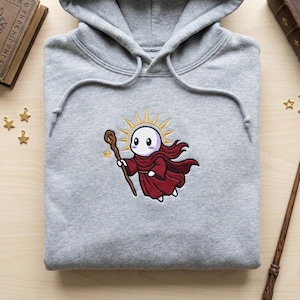 Könnte beinhalten: Grauer Kapuzenpullover mit einem gestickten Cartoon-Charakter mit roter Robe und Sonnenhalo, der einen Stab hält. Der Hoodie ist sauber auf einer Holzoberfläche gefaltet. Ein skurriles Design.