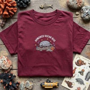 May include: A maroon t-shirt featuring a central embroidered graphic of a grey isopod on rocks with the text "ARMORED OCEAN BUG" arched above it. The design includes small bubbles, yellow specks, and orange coral accents surrounding the creature.