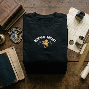May include: Black crew neck sweatshirt featuring the embroidered text "Riders Quadrant" above a gold dragon and blue stars. The image includes an Academy patch and antique books on a wooden surface. The sweatshirt is folded.