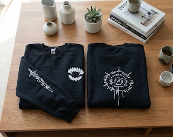 May include: Two black sweatshirts with white graphic designs are displayed on a wooden table. One sweatshirt features a barbed wire design on the sleeve and a toothy grin on the chest. The other has a circular, abstract design.