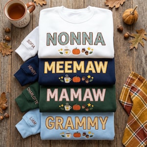 May include: A stack of four crewneck sweatshirts in white, navy blue, dark green, and light blue. Each features embroidered text reading NONNA, MEEMAW, MAMAW, or GRAMMY, with autumn-themed icons of a coffee mug, pumpkin, and acorns. Sleeves show names.