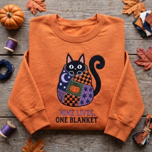 May include: An orange crewneck sweatshirt featuring an embroidered black cat wrapped in a patchwork blanket with pumpkin, star, and moon patterns. Below the cat, the text reads NINE LIVES, ONE BLANKET in dark blue, sans-serif capital letters.