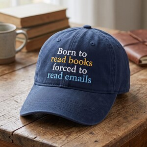May include: A navy blue baseball cap featuring embroidered text that reads Born to read books in white and yellow, followed by forced to read emails in white and light blue. The cap has a curved brim and a structured crown with ventilation eyelets.