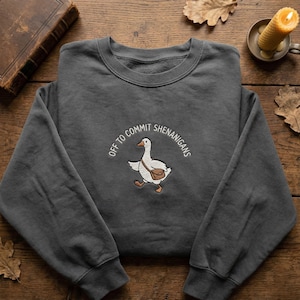 May include: Charcoal grey sweatshirt featuring an embroidered goose with a satchel, and the text "OFF TO COMMIT SHENANIGANS" in an arc. The goose is walking. The sweatshirt is folded on a wooden surface, with a candle and book.