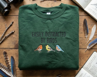 May include: A dark green sweatshirt with the embroidered phrase "EASILY DISTRACTED BY BIRDS" above three embroidered birds. The birds are a robin, a yellow finch, and a bluebird. The sweatshirt is on a wooden surface with birdwatching accessories.
