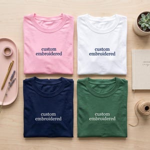 May include: Four t-shirts in pink, white, navy blue, and green, each with the words "custom embroidered" in a contrasting color. The shirts are neatly folded on a light wooden surface, with stationery and a small succulent plant nearby.