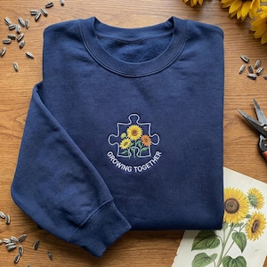 Peut inclure: Un sweat-shirt bleu marine avec un col rond. Le sweat-shirt présente un motif brodé de tournesols à l'intérieur d'une pièce de puzzle, avec les mots "Growing Together" en dessous. Le sweat-shirt est fait d'une matière douce.