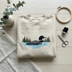 May include: A beige sweatshirt with an embroidered design of a loon swimming in blue water with green trees in the background. The loon has black and white markings. A wooden embroidery hoop, scissors, and thread are also visible.