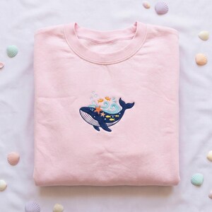May include: A light pink sweatshirt with an embroidered design of a navy blue whale with a white belly, surrounded by orange starfish, yellow fish, and blue waves. The sweatshirt is folded neatly.