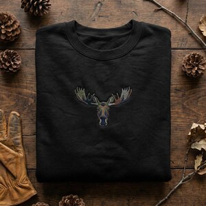 May include: Black sweatshirt featuring an embroidered moose head with antlers. The moose design incorporates shades of green, brown, and blue. The sweatshirt is folded and placed on a wooden surface, with pine cones and leaves around it.