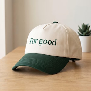 May include: A baseball cap with an off-white crown and a dark green brim. The front of the cap features the embroidered text "For good" in a dark green serif font. The cap has a structured five-panel design with a matching dark green button on top.