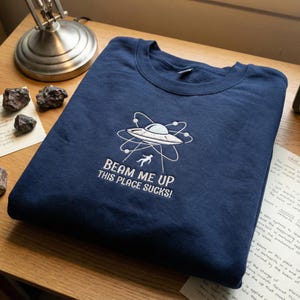 May include: A navy blue sweatshirt featuring a white embroidered design of a UFO, atom, and a person being beamed up. The text "BEAM ME UP THIS PLACE SUCKS!" is below the graphic. The sweatshirt is folded on a wooden surface.