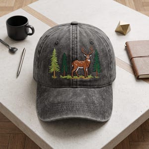 May include: A charcoal grey baseball cap with a distressed finish. The front panel features an embroidered graphic of a brown deer with antlers standing in a grassy area between three green pine trees of varying shades and sizes.