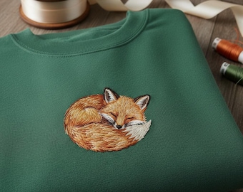 May include: A forest green sweatshirt with an embroidered sleeping fox design. The fox is depicted in shades of orange, brown, and white, curled up in a ball. The sweatshirt is made of a soft material and is a crew neck style.