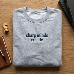 May include: A heather gray sweatshirt with the embroidered text "sharp minds collide" in navy blue and yellow. The sweatshirt is folded on a wooden surface, with a chess piece and a notebook visible.