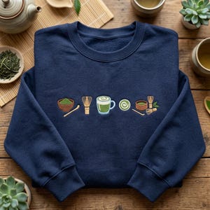 May include: Navy blue sweatshirt featuring embroidered matcha tea elements: a bowl of matcha, a whisk, a mug of matcha, a matcha swirl, and a bowl with a whisk. The sweatshirt is on a wooden surface with tea-related items.