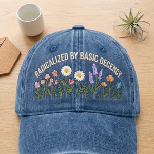 May include: A blue denim baseball cap featuring embroidered wildflowers in shades of pink, white, orange, and purple. The text RADICALIZED BY BASIC DECENCY is stitched in a cream-coloured font above the floral design on the front panel.