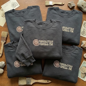 May include: Five charcoal gray sweatshirts with the text "ABSOLUTELY SHELL-AR" and a seashell design embroidered in tan. The sweatshirts are arranged on a wooden surface with postcards, paintbrushes, and rocks.