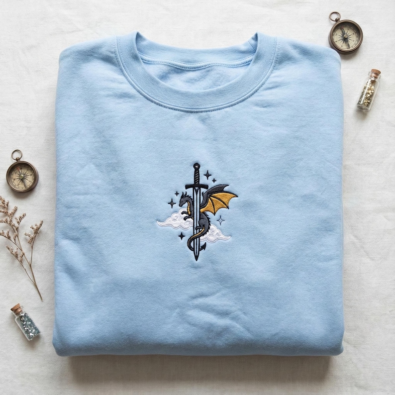 May include: A light blue sweatshirt with an embroidered design featuring a dragon, sword, and stars. The dragon has yellow wings and is positioned on a cloud. The sweatshirt is folded neatly on a white surface.