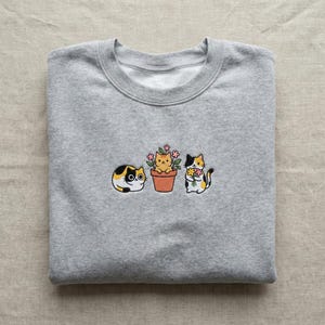 May include: A gray sweatshirt with an embroidered design of three cartoon cats. One cat is lying down, another is in a flower pot, and the third is holding flowers. The cats are in shades of yellow, black, and white.