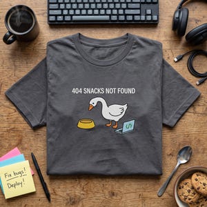May include: A dark grey t-shirt featuring a graphic of a white goose with an orange beak standing next to a yellow bowl and a small open laptop. The text 404 SNACKS NOT FOUND is printed in white above the goose illustration on the centre chest.
