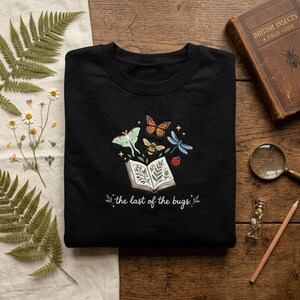 Könnte beinhalten: Ein schwarzes Sweatshirt mit Rundhalsausschnitt und einer Stickerei, die ein offenes Buch mit botanischen Illustrationen zeigt, umgeben von einem Mondfalter, Monarchfalter, einer Biene, Libelle und einem Marienkäfer, mit dem Text "the last of the bugs."