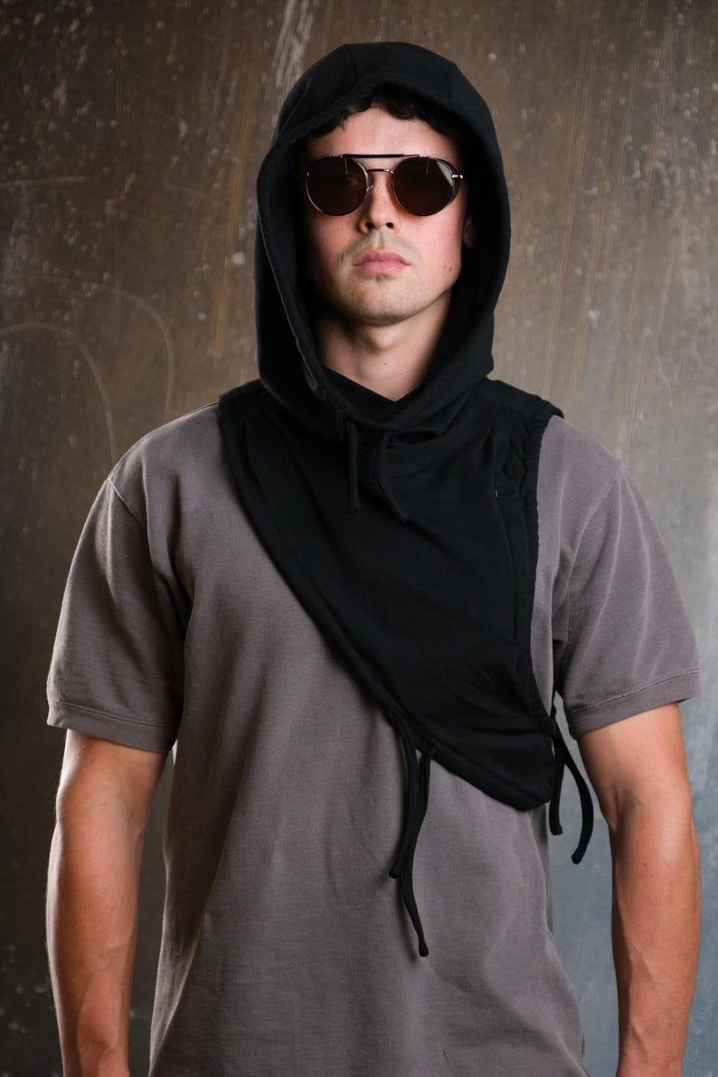 Op de afbeelding: Een zwarte sjaal met capuchon en trekkoorden, gedragen over een grijs t-shirt. De sjaal heeft een uniek asymmetrisch ontwerp. De persoon draagt ​​aviator zonnebrillen. De achtergrond is een gevlekt grijs en bruin.