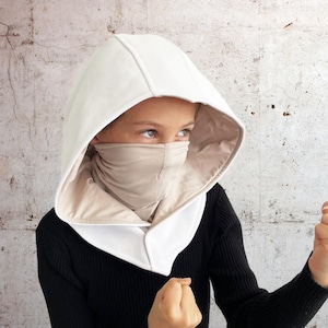 Peut inclure: Une cagoule blanche à capuche avec une doublure beige. La cagoule est tirée sur la tête d'une personne portant une chemise noire. La personne regarde vers la droite et a les poings serrés.