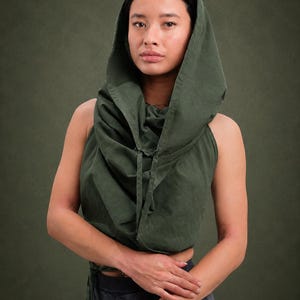 May include: A sleeveless olive green crop top featuring an attached hood and a draped cowl neckline. The garment includes thin fabric ties at the front and is constructed from a solid-coloured textile with visible seams along the hood and bodice.