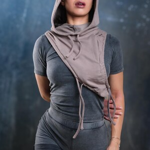 Op de afbeelding: Een grijze hoodie met col en bijpassende shorts. De top heeft een trekkoord en een ritssluiting. Het model draagt een zonnebril. De outfit is een casual streetwear-stijl.