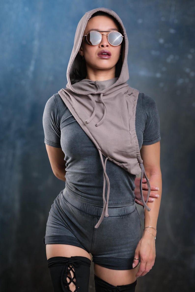 Op de afbeelding: Een grijze hoodie met col en bijpassende shorts. De top heeft een trekkoord en een ritssluiting. Het model draagt een zonnebril. De outfit is een casual streetwear-stijl.
