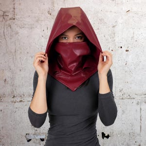 May include: A deep red faux leather hood and face mask set. The hood features a pointed top and covers the head, while the matching face mask covers the nose and mouth area, extending down to a point at the chest, creating a cohesive head covering.