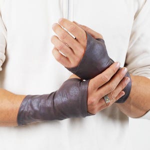Puede incluir: Un par de guantes sin dedos de cuero marrón con un acabado texturizado. Los guantes se extienden hasta la muñeca y tienen una costura en la parte posterior de la mano.