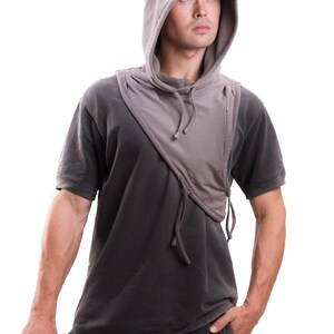 Op de afbeelding: Een grijze sjaal met capuchon en trekkoord, gedragen over een donkergrijs t-shirt. De sjaal heeft een uniek asymmetrisch ontwerp. De persoon houdt een zonnebril vast en heeft zijn hand in zijn zak.