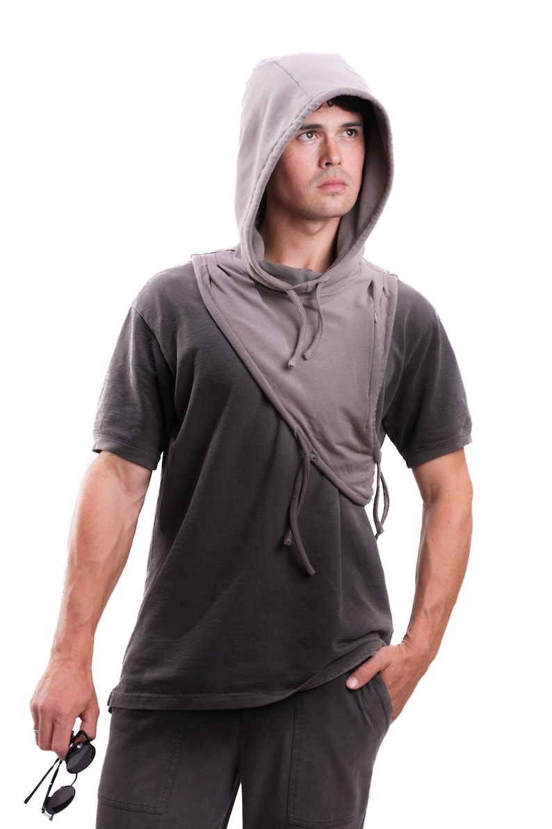 Op de afbeelding: Een grijze sjaal met capuchon en trekkoord, gedragen over een donkergrijs t-shirt. De sjaal heeft een uniek asymmetrisch ontwerp. De persoon houdt een zonnebril vast en heeft zijn hand in zijn zak.