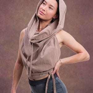 May include: A sleeveless taupe hooded top featuring a draped cowl neckline, a drawstring hood, and wrap-around waist ties. The garment is paired with dark grey shorts that have a wide, ribbed elastic waistband and a cuffed hemline.