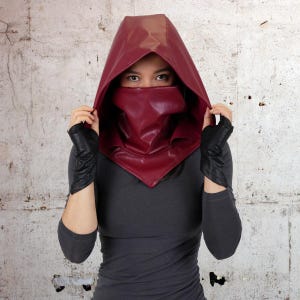 May include: A woman wearing a burgundy leather hooded cowl with a face covering. The cowl is pulled up over her head and covers her face, leaving only her eyes visible. She is wearing a dark grey long-sleeved shirt.