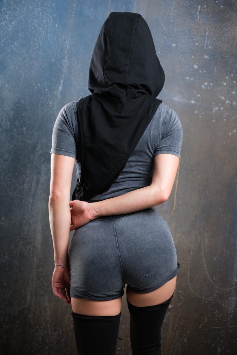 Op de afbeelding: Een persoon draagt een grijs t-shirt, een korte broek en een zwarte capuchon. De persoon staat met de rug naar de camera, de armen gekruist achter de rug. De outfit bevat zwarte kniehoge sokken.