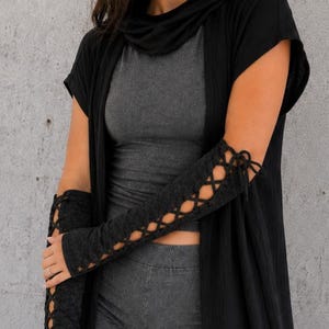K&ouml;nnte beinhalten: Ein &auml;rmelloses Oberteil in Dunkelgrau und eine passende Shorts, kombiniert mit einer langen, vorne offenen schwarzen Strickjacke. Das Outfit umfasst schwarze Armstulpen und passende Beinstulpen, beide mit Schn&uuml;rdesign und schwarzen B&auml;ndern.