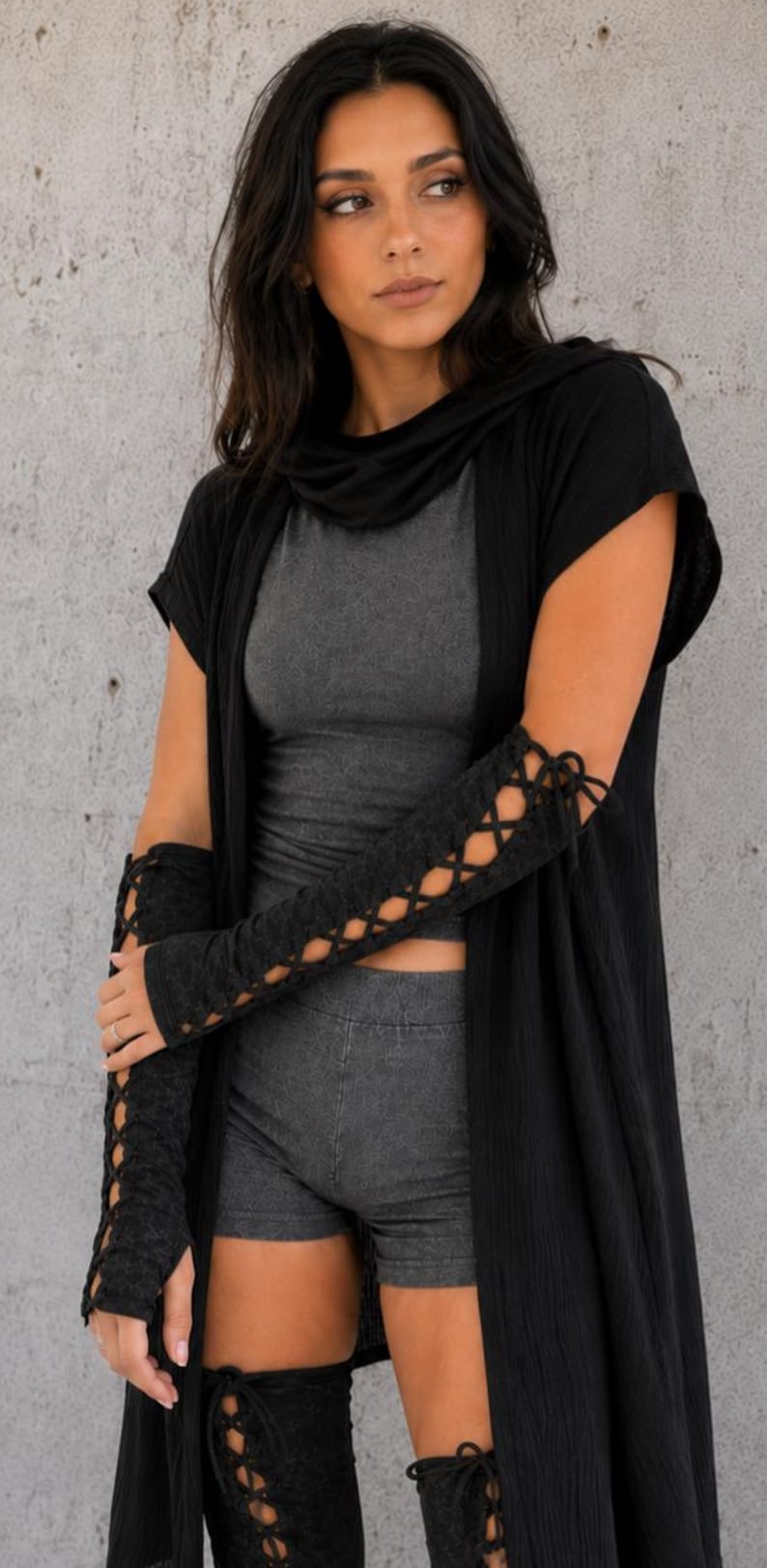 K&ouml;nnte beinhalten: Ein &auml;rmelloses Oberteil in Dunkelgrau und eine passende Shorts, kombiniert mit einer langen, vorne offenen schwarzen Strickjacke. Das Outfit umfasst schwarze Armstulpen und passende Beinstulpen, beide mit Schn&uuml;rdesign und schwarzen B&auml;ndern.