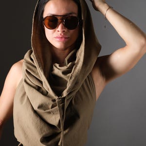 Könnte beinhalten: Olivgrünes, ärmelloses Crop-Top mit drapierter Kapuze und Kordelzug. Das Model trägt braune Pilotenbrillen. Das Oberteil wird mit dunkler Hose kombiniert.