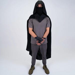 May include: A person wearing a black hooded cloak with a black face mask. The person is wearing a grey t-shirt and grey trousers. The person is standing on a white background.