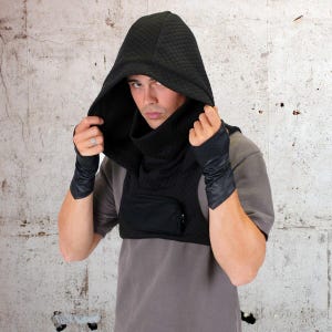 May include: A black hooded garment with a quilted texture and a zippered pocket on the front. The hood is pulled up over the head, and the garment is worn over a gray shirt.