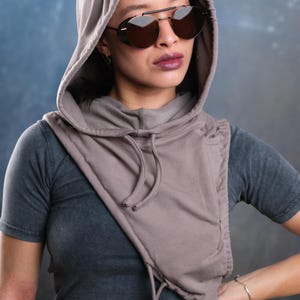 Op de afbeelding: Een capuchonsjaal in een gedempte taupe kleur, gecombineerd met een donkergrijs t-shirt. De sjaal heeft een trekkoord en valt over de schouders. De persoon draagt een donkere zonnebril met een zilveren montuur.