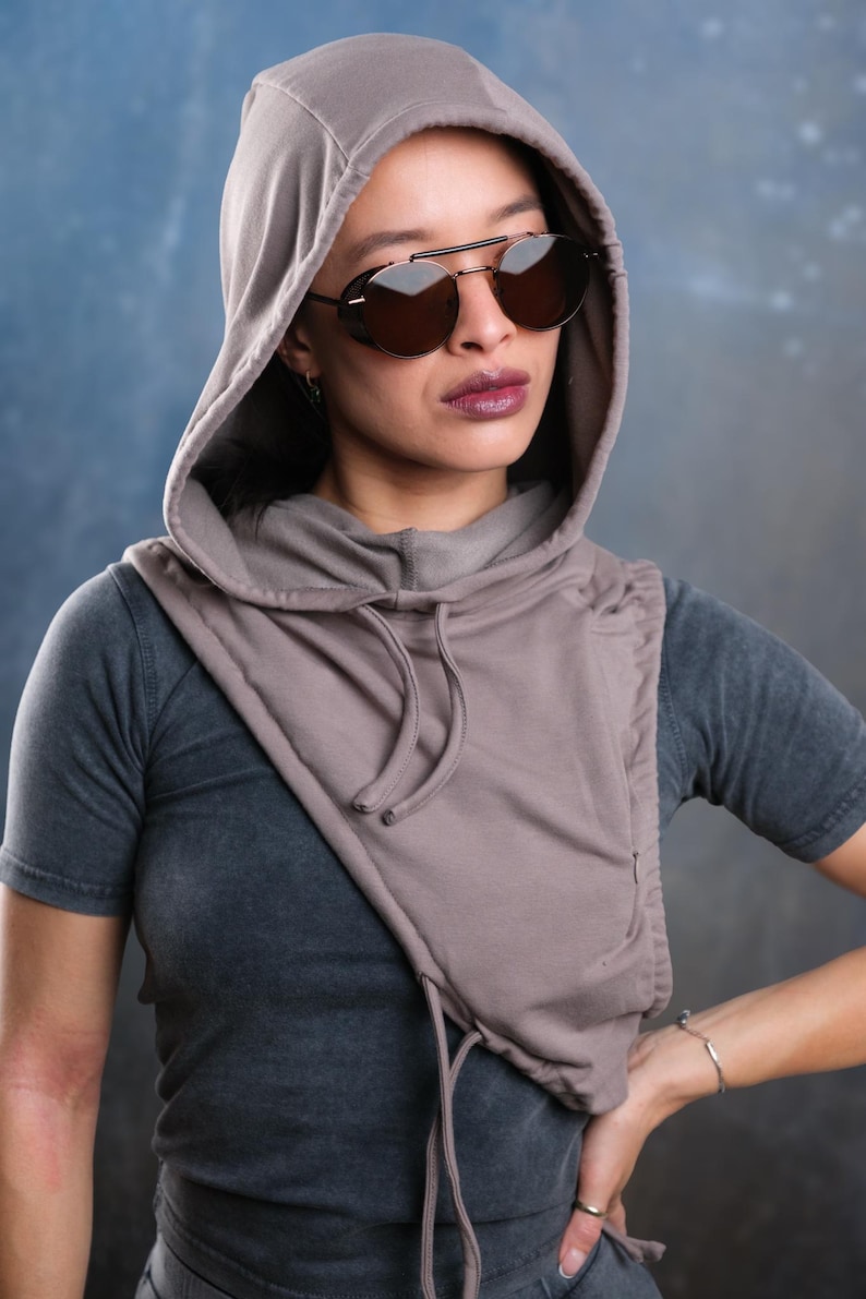 Op de afbeelding: Een capuchonsjaal in een gedempte taupe kleur, gecombineerd met een donkergrijs t-shirt. De sjaal heeft een trekkoord en valt over de schouders. De persoon draagt een donkere zonnebril met een zilveren montuur.