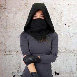 May include: A black hooded scarf with a quilted pattern covers the face of a person wearing a gray long-sleeved shirt. The person is standing in front of a concrete wall.