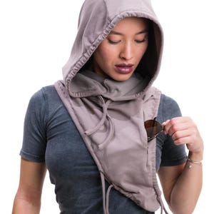 Op de afbeelding: Een kap sjaal in een gedempte mauve kleur, met een ingebouwde zak. De sjaal heeft trekkoorden en wordt over een grijs t-shirt gedragen. Zonnebrillen worden in de zak gestopt. Het ontwerp is casual en praktisch.
